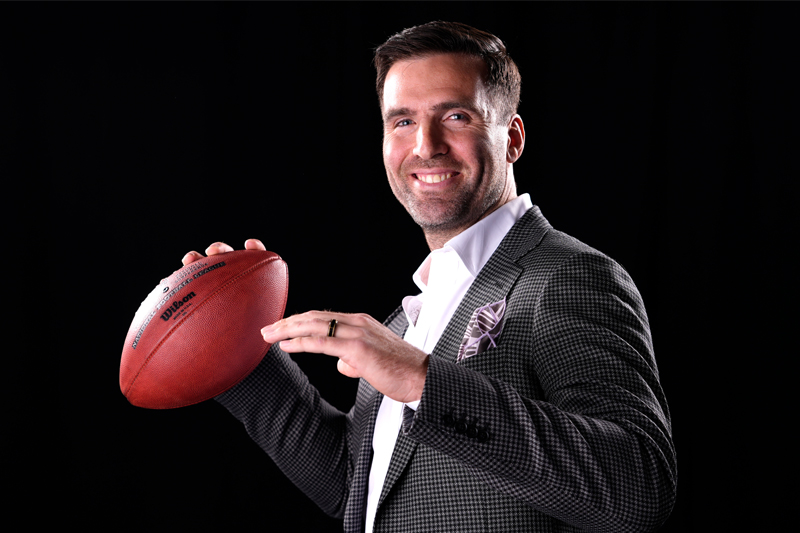 NFL Quarterback, Joe Flacco, poses with a football as if he's about to throw it, and smiles for the camera.