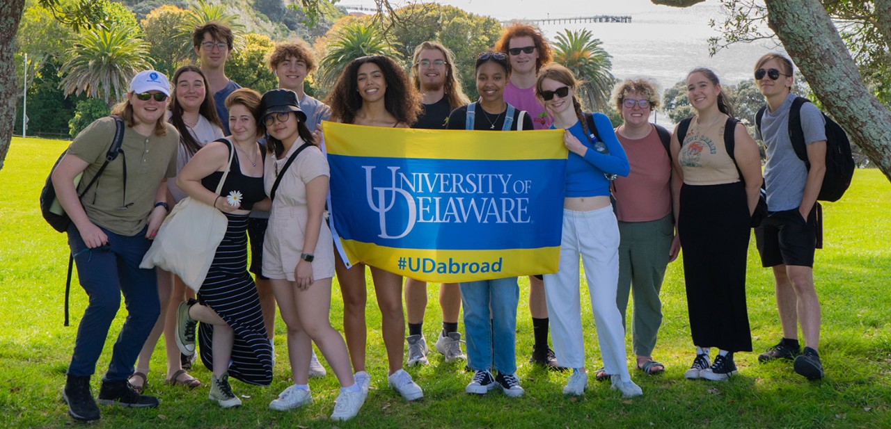 A group of University of Delaware students taking a picture during a study abroad trip in Auckland, New Zealand. They are holding a University of Delaware flag with a hashtag that says "#UDabroad" on it.