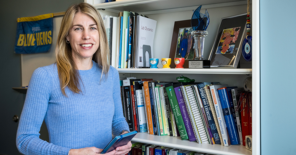 Kinesiology and Applied Physiology Professor Shannon Lennon stands near a full bookcase in her office wearing a blue sweater. She was recently award the Torch Award from the UD Women's Caucus for advancing women's equity on campus.