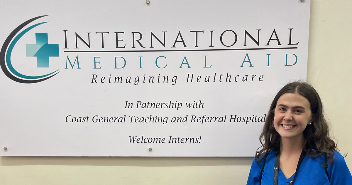 Senior nutrition and dietics major Katie Hamelin poses in scrubs next to the sign for International Medical Aid, where she took part in a rare nutrition-focused internship in Kenya. 