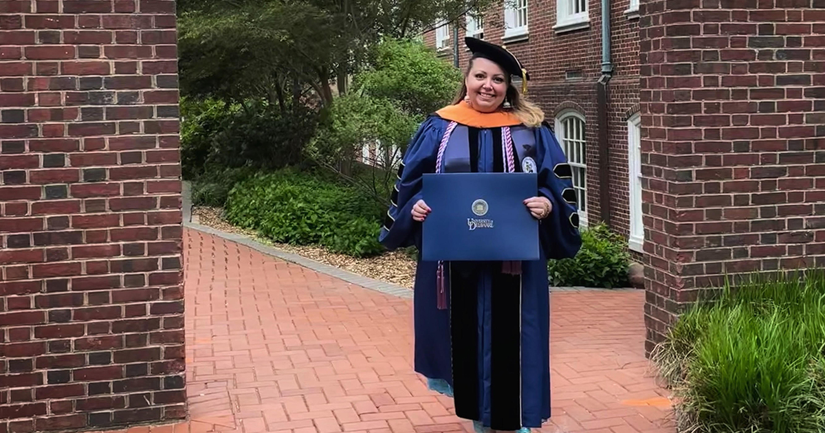 Rebecca Ivory stands on UD's Campus in her cap and gown after graduation, holding up her Doctor of Nursing Practice degree.