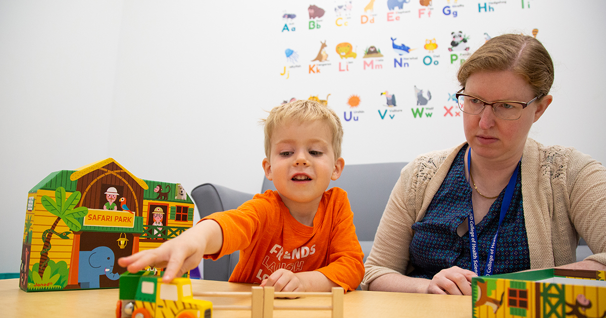 A boy in a bright orange shirt smiles as he plays with toy truck that's part of a colorful Safari Park toy while Amanda Van Owen Horne observes. The professor of communication and sciences disorders is using play as a form of intensive language development therapy.  