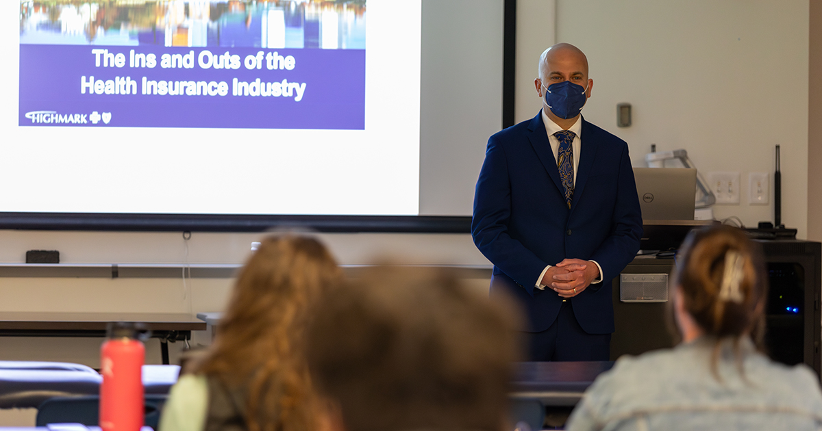 Highmark president Nick Moriello wears a mask and a suit and tie while talking to a biomechanics and movement science graduate-level class in the Health Sciences Complex. Behind him is a Powerpoint on a slide titled "The  Ins and Outs if the Health Insurance Industr.y."
