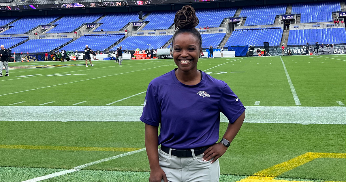 Asha Rogers poses in a purple Baltimore Ravens shirt with her hand on her hip on the field of M&T Bank Stadium, home of the Baltimore Ravens. She completed a clinical rotation with the team before graduating with her doctor of physical therapy.