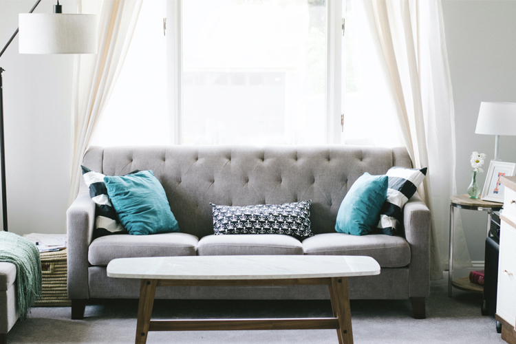 a gray couch with blue cushions in front of a window with sheer curtains