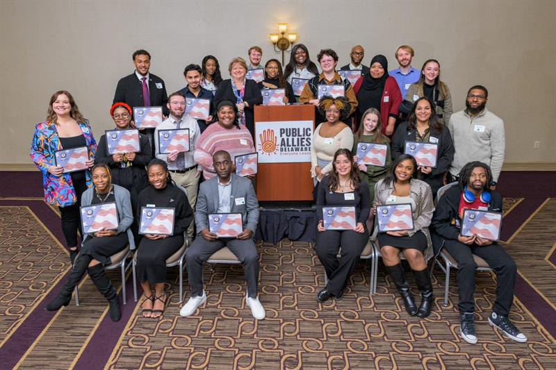 The 2022-23 Public Allies cohort stands huddled together smiling in a group photo while dressed in professional attire at their AmeriCorps induction ceremony