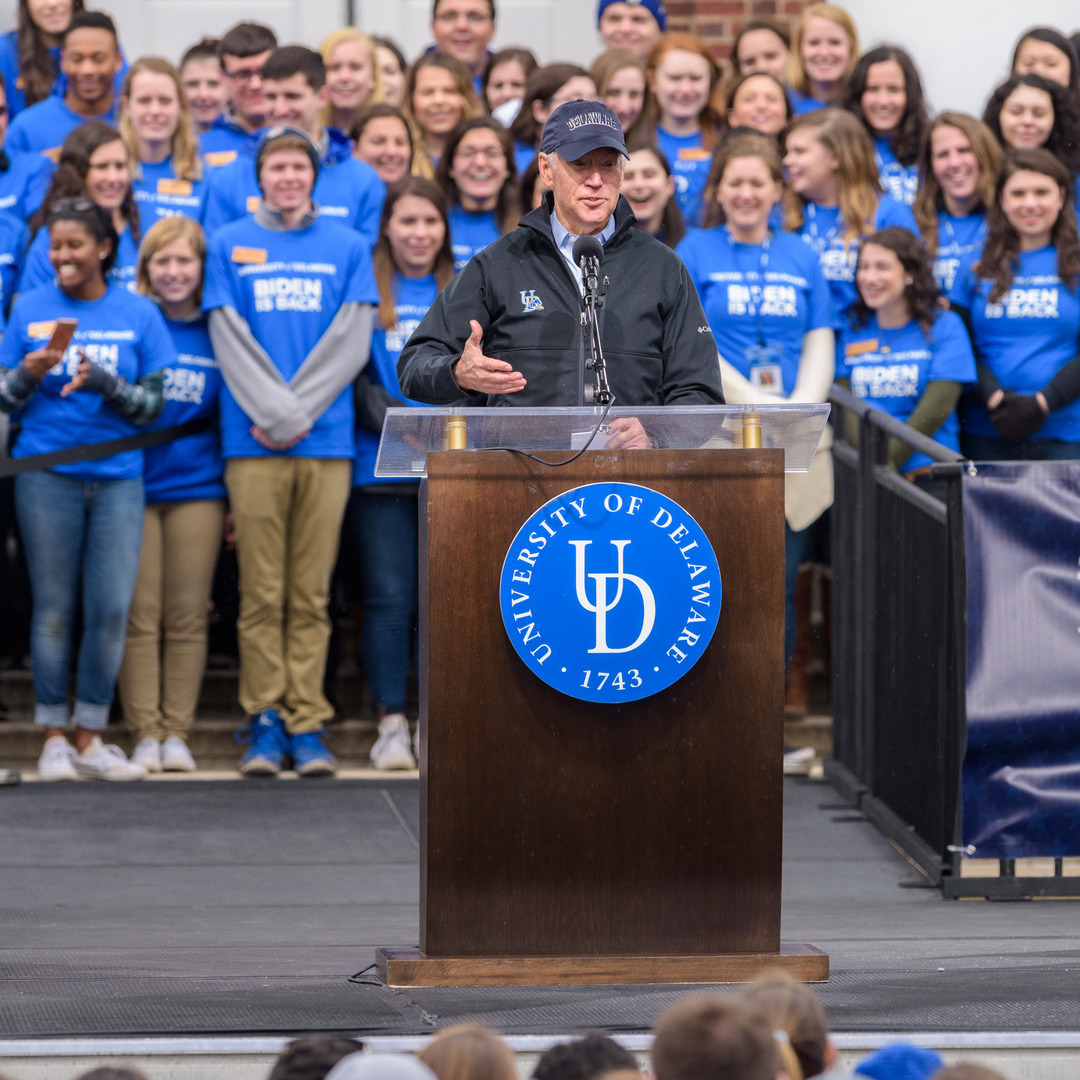 Photo of President Joe Biden stands in front of a podium bearing the University of Delaware monogram, a large crowd of students is gathered behind him.