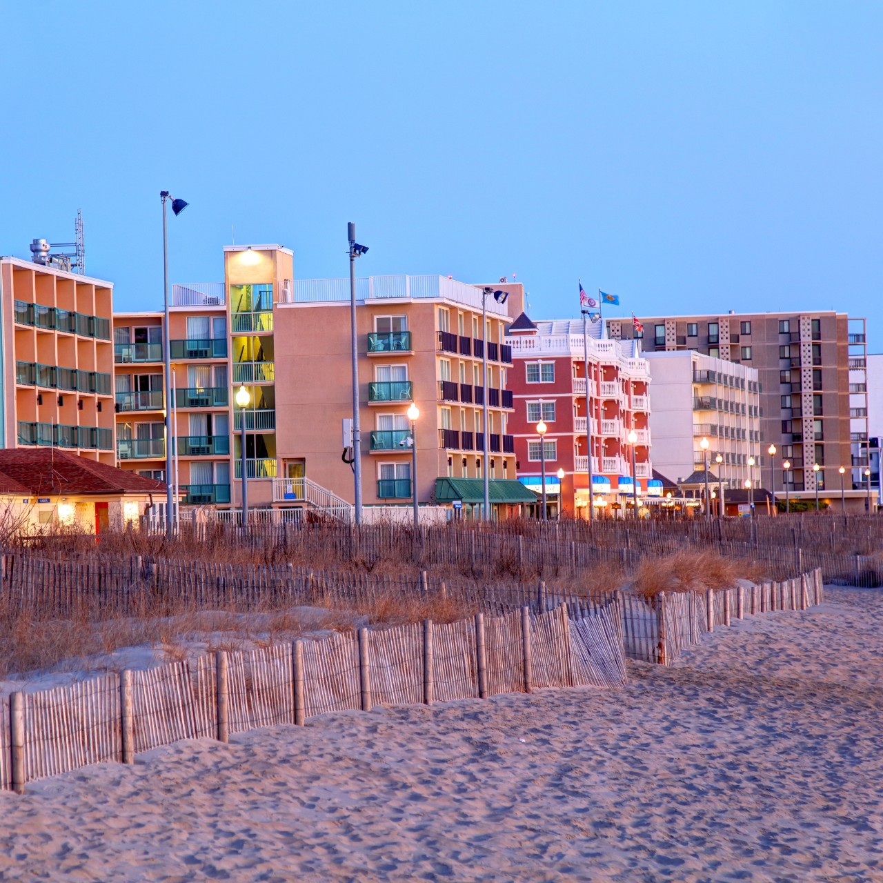 A photo of colorful buildings on the Rehoboth Beach, Delaware boardwalk at sunset with sand and dunes in the foreground.