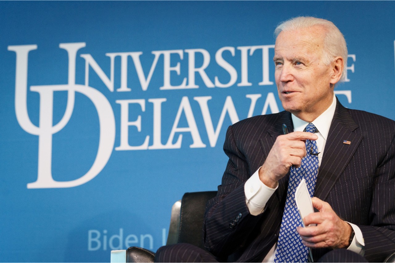 Photo of President Joe Biden wearing a suit and tie and lapel microphone while speaking onstage. A blue backdrop with the white University of Delaware logo sits behind him in the background.