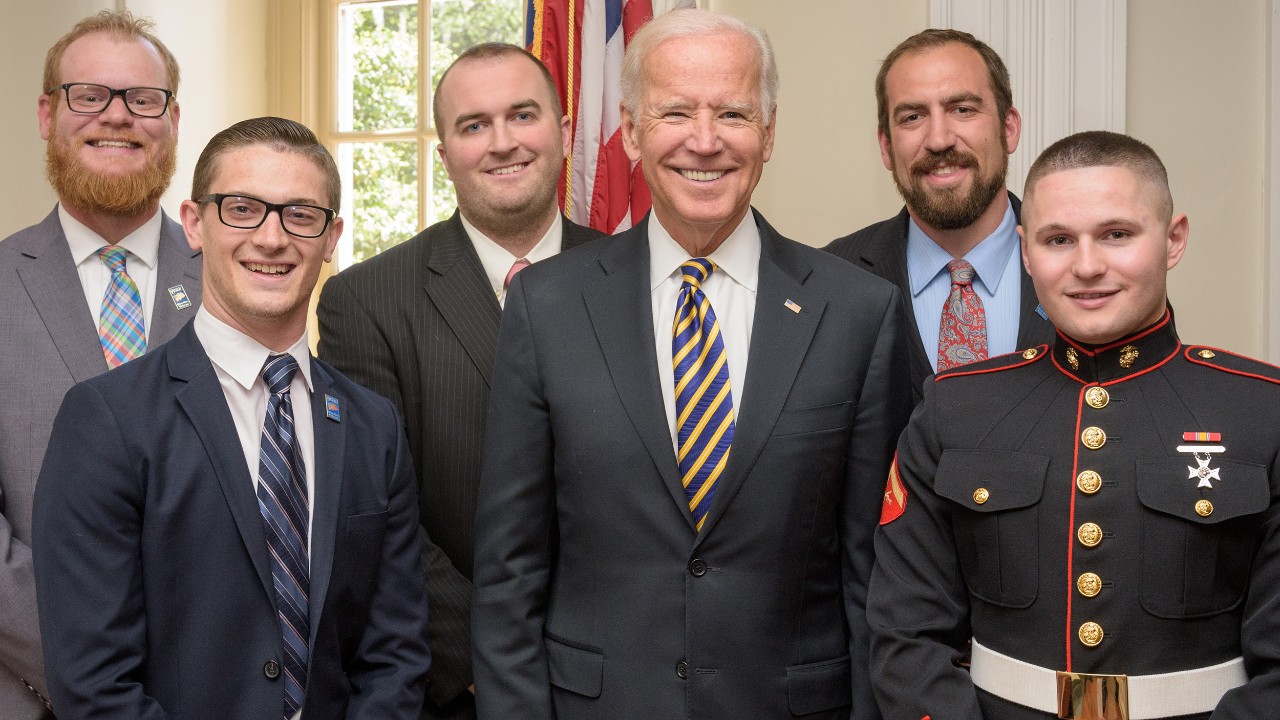 Photo of President Joe Biden smiling while gathered with members of Blue Hen Veterans dressed in suits and military uniforms