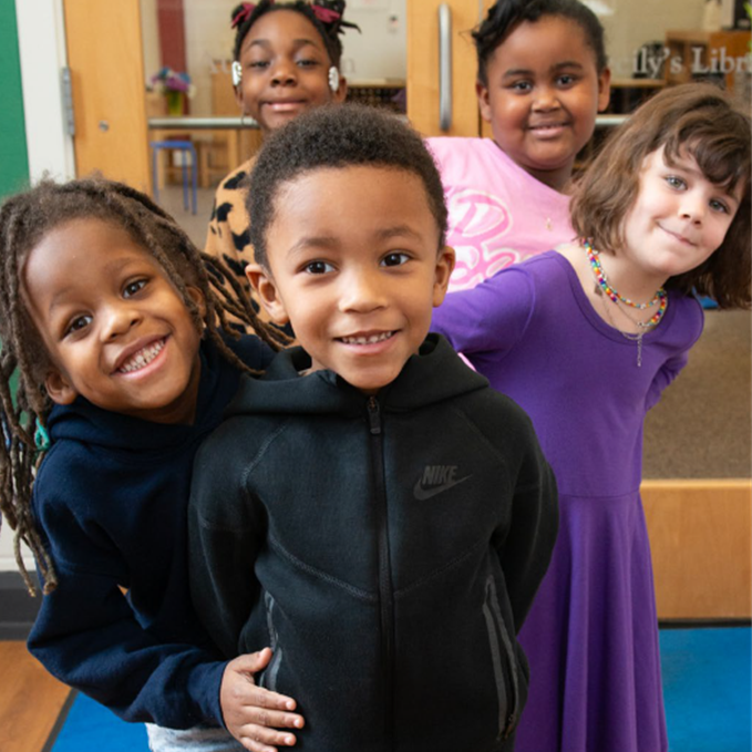 Photo of five young children standing smiling and giggling behind each other in a line peaking out on different sides