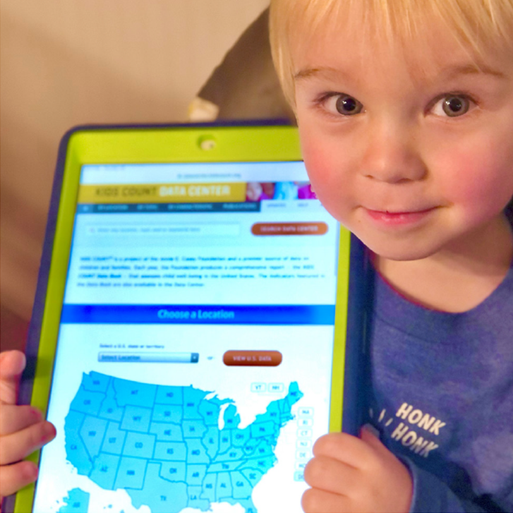 Photo of a small child smiling at the camera while holding a tablet displaying the KIDS COUNT Data Center website