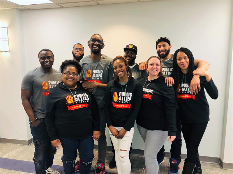 Photo of a previous Public Allies Delaware cohort gathered close together and smiling while wearing black and grey shirts with the Publlic Allies branding displayed