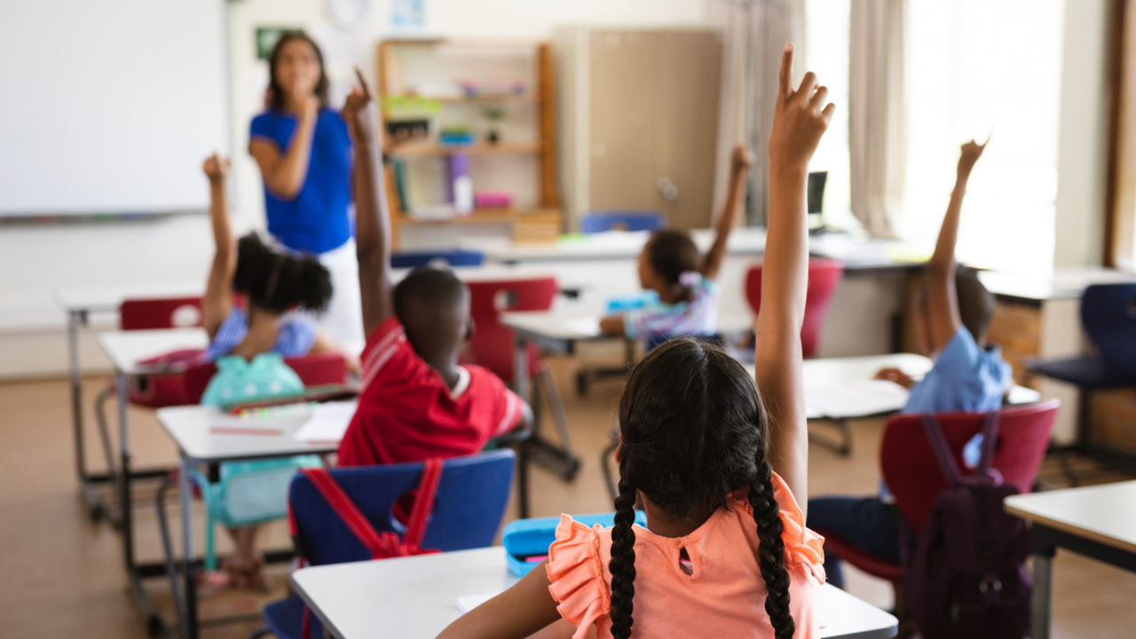 Stock photo of elementary school students raising hands in classroom, with teacher standing at the front