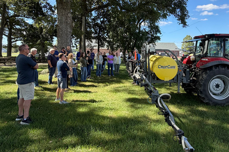 A group gathers under shade trees for an outdoor demonstration beside a red tractor equipped with a yellow agricultural sprayer.