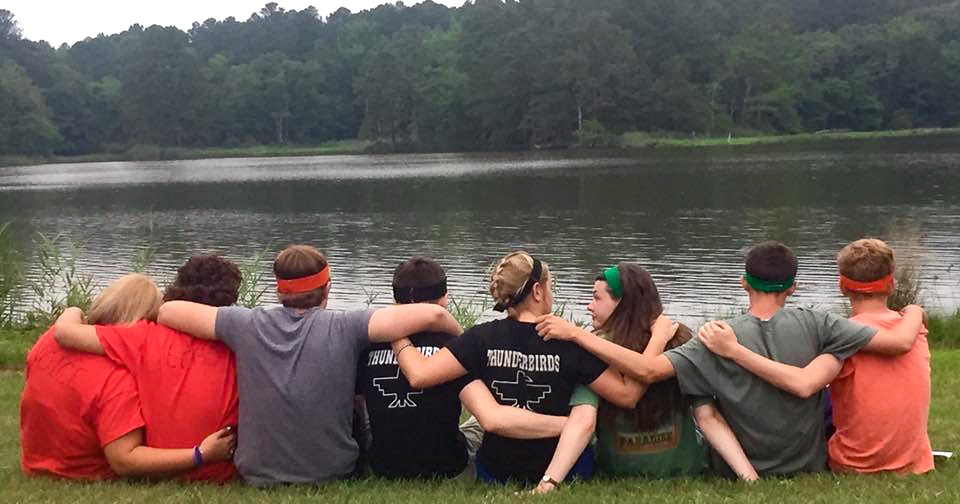 eight kids sitting with their arms around each other looking at a lake