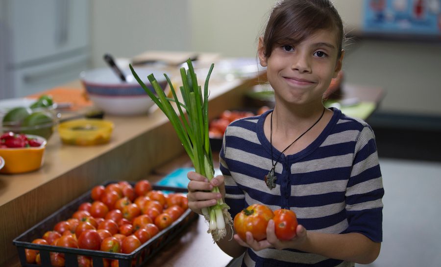 A young girl holds vegetables in a kitchen.