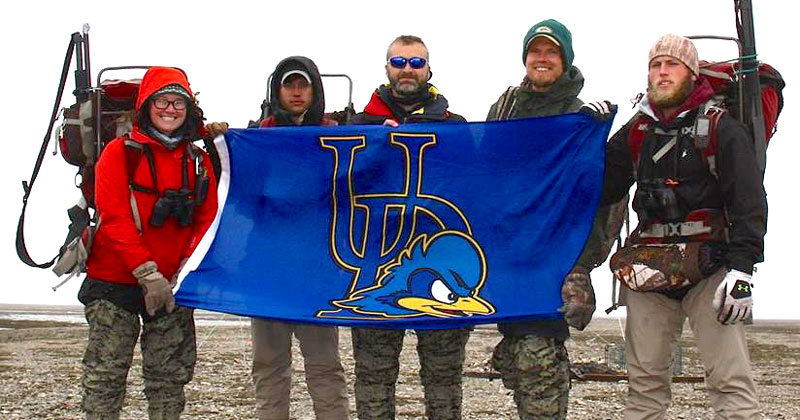 A photo of professor Chris Williams with students holding a UD Bluehen flag
