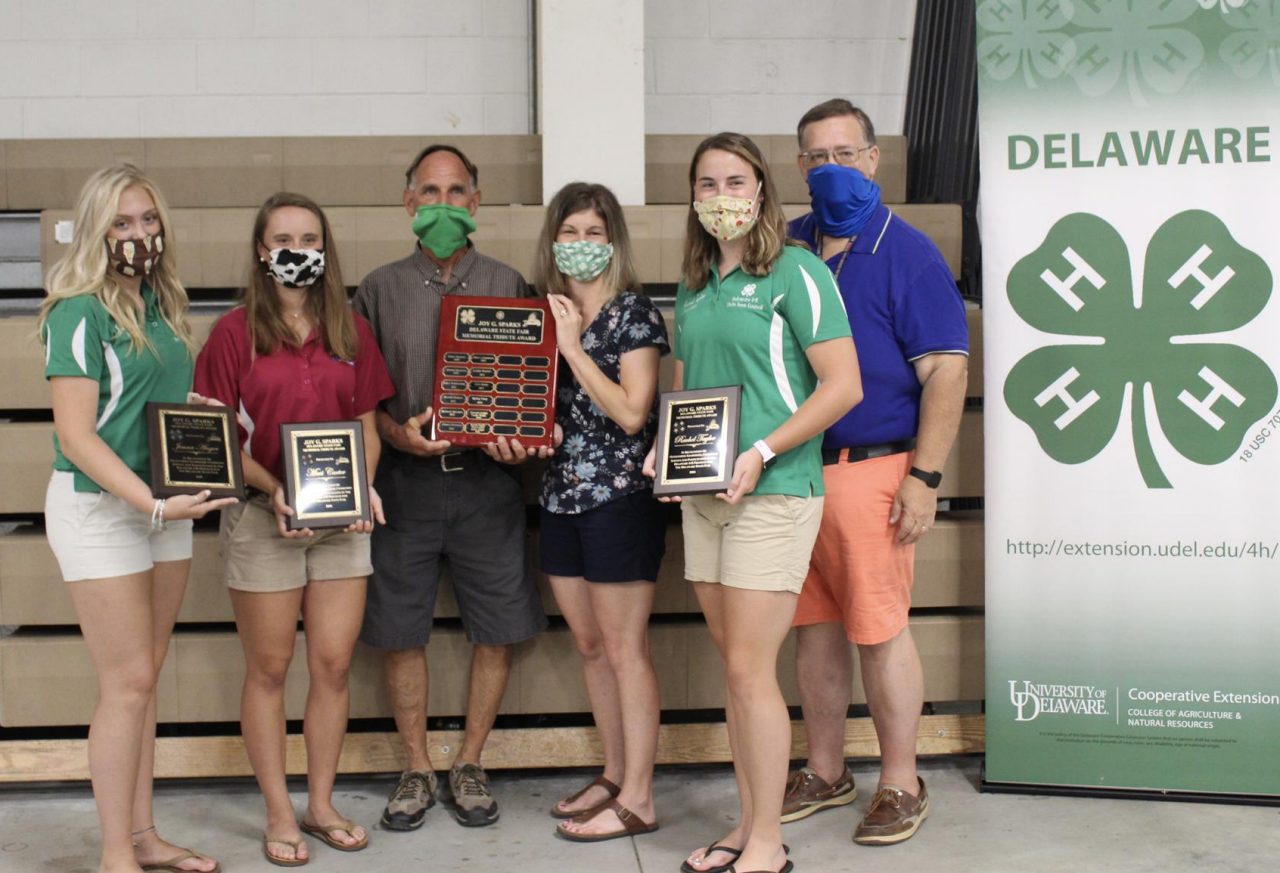 Jenna Anger, Maci Carter and Rachel Taylor, pose (with COVID masks) with Alex Gooden and Lindsay Hughes, representing the family of Joy Sparks, and Doug Crouse, State 4-H Program Leader. Each hold their received award plaque.