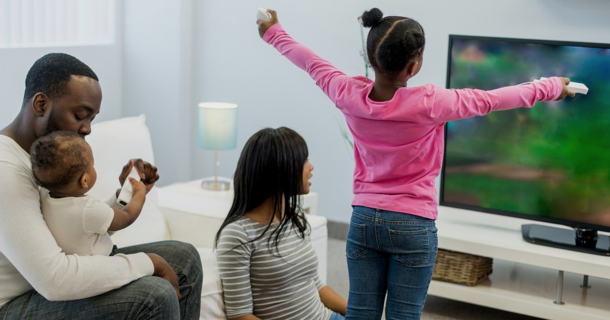 A young girl stands and plays an active video game, her arms stretched out wide, while her family watches.