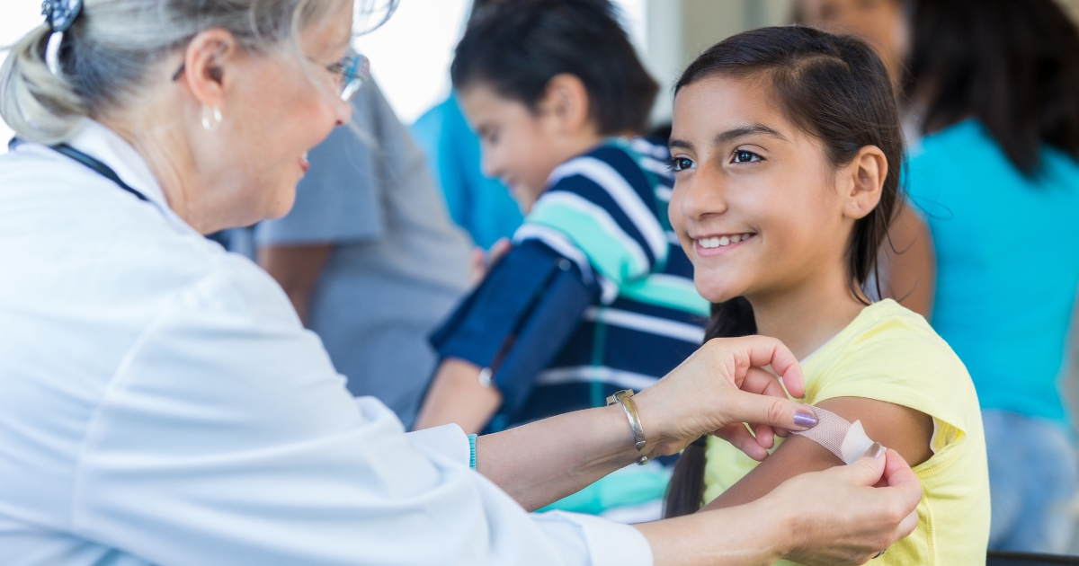 flu_vaccine_website_properties_img_extension_1200x630 - 1 A smiling nurse puts a bandaid on a girl after vaccination.