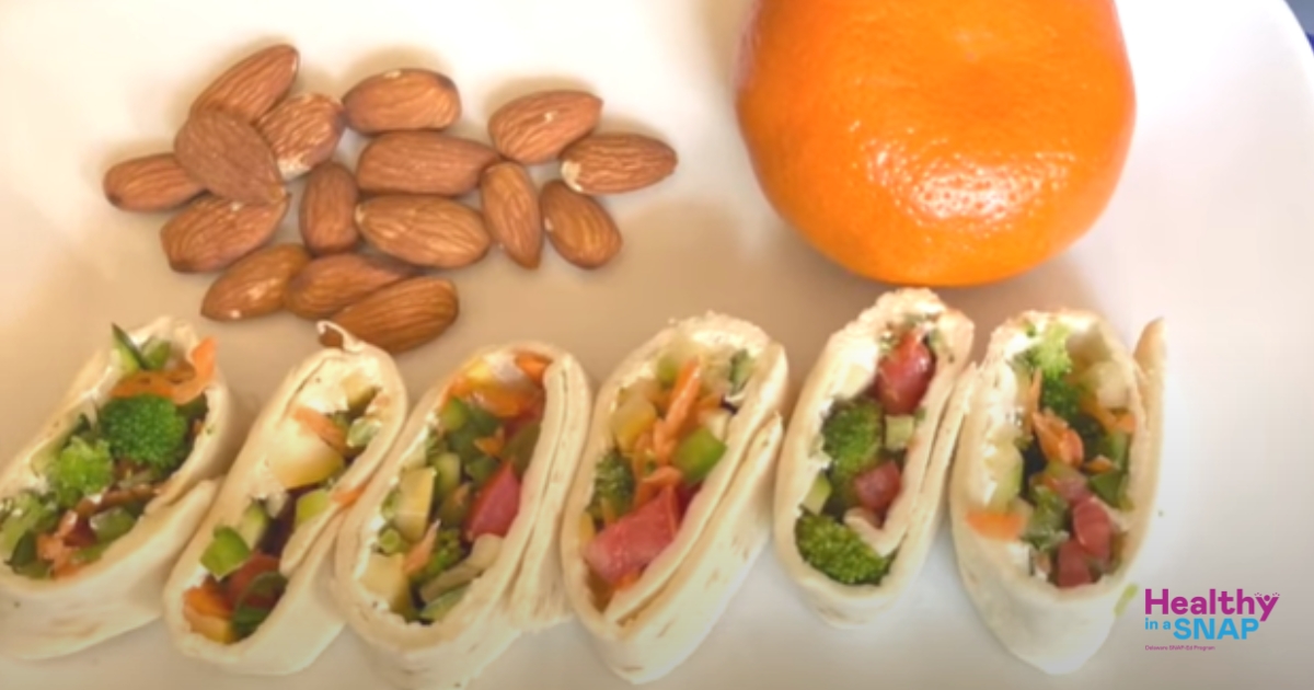 A photo of 6 sliced zucchini wraps in a row next to an orange and nuts