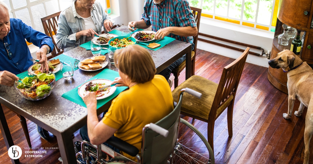 Four older people, one in a wheelchair sharing a meal around a table and a dog watching Four older people, one in a wheelchair sharing a meal around a table and a dog watching