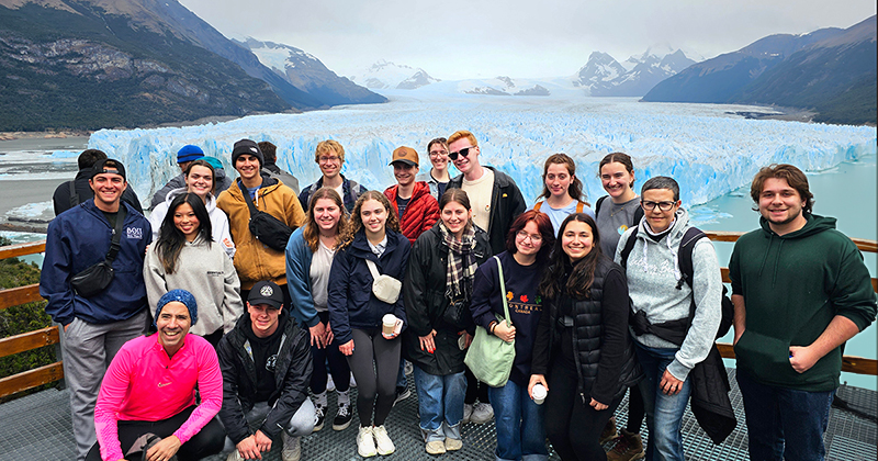 A group of smiling University of Delaware students, faculty and tour guides in front of a large ice field  surrounded by mountains.