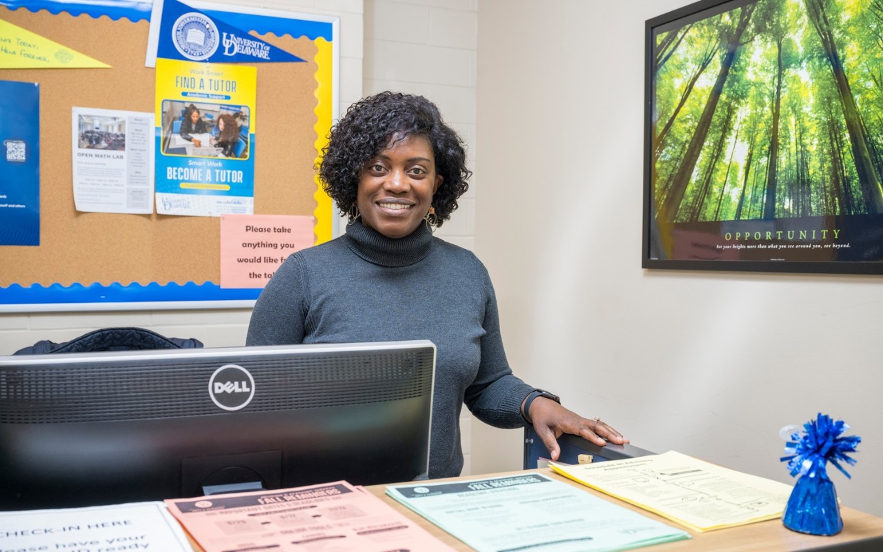 A Student Success Center staff person stands behind a counter with a computer monitor, flyers and posters, smiling and welcoming students.
