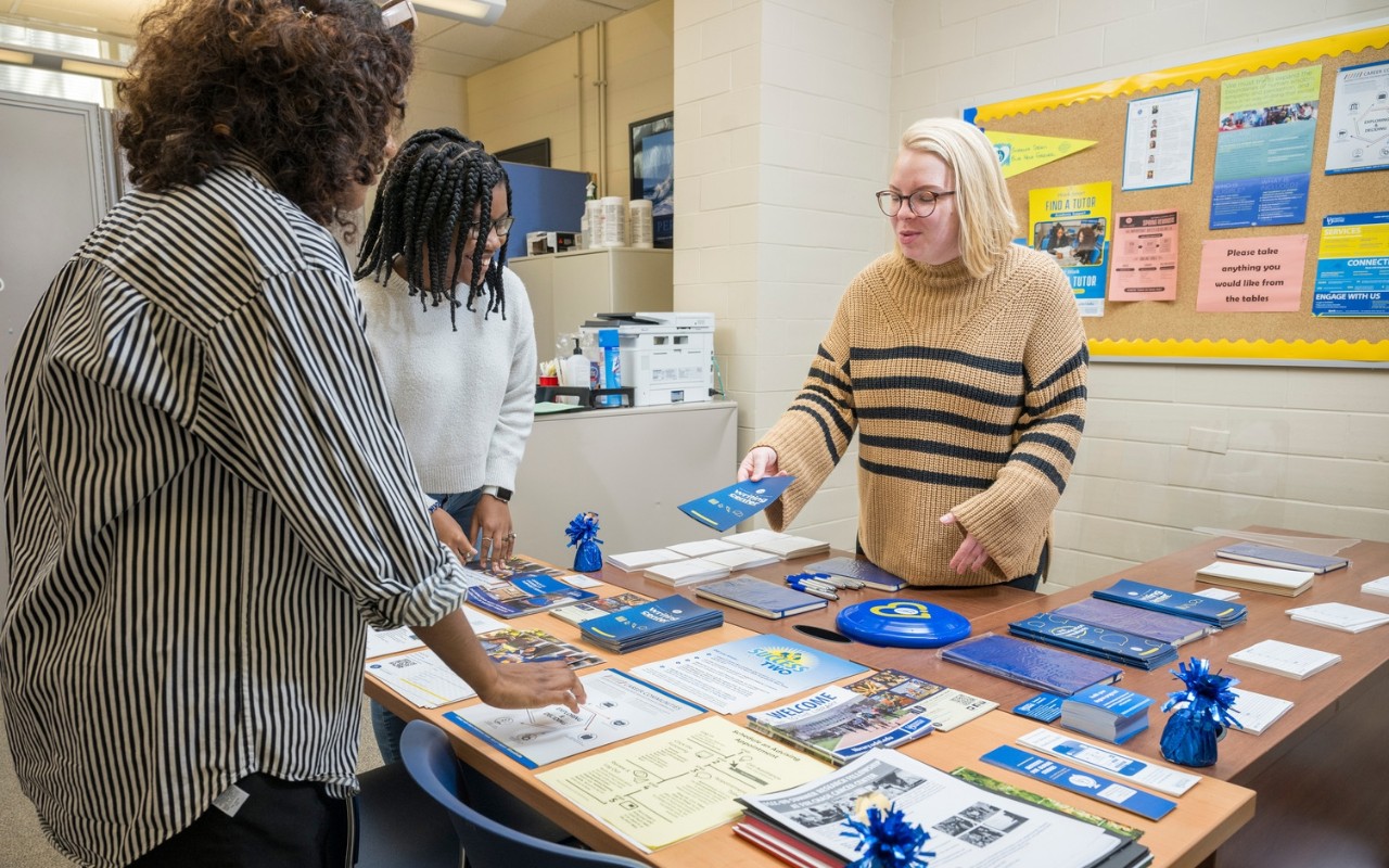 A Student Success Center staff person standing inside an office behind a table with brochures and flyers talks to two University of Delaware students and hands one of them a pamphlet.