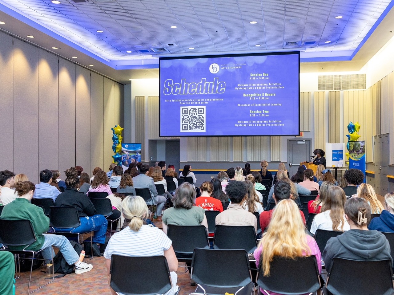 A group of students sitting in the Trabant Multipurpose Room listen to a presentation with slides on a large screen.