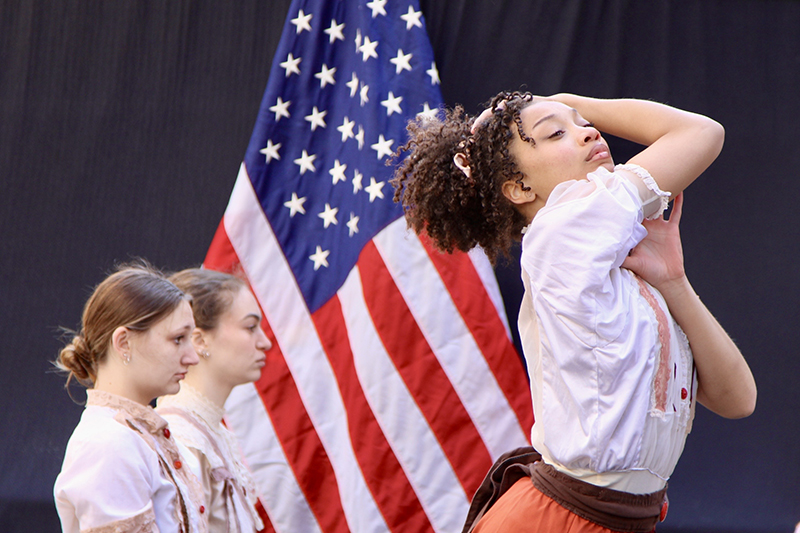 Recent graduate Marissa Jackson, public policy major with a dance minor, performs an excerpt from Project 146 at the 114th anniversary Triangle Fire Commemoration. 3 girls dancing in front of American flag