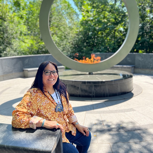 Rachelle Pablo sits on a bench on a stone patio with a large contemporary sculpture of a circle behind her.