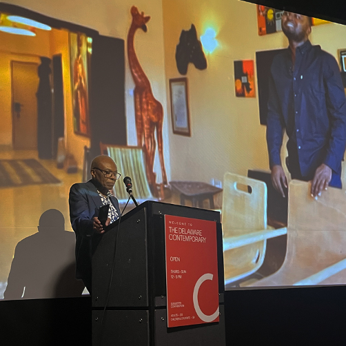 Ikem Okoye delivers a presentation while standing behind a podium with a microphone and slideshow on a screen behind him.