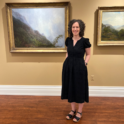 A smiling woman wearing a black dress stands inside a museum in front of two paintings.