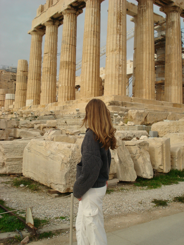 The back of a woman with long brown hair, Riley Morrison, looking out over scenery in Athens, Greece