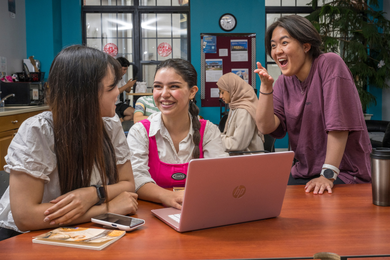 Three international female students smiling and talking at a table with a laptop and notebooks