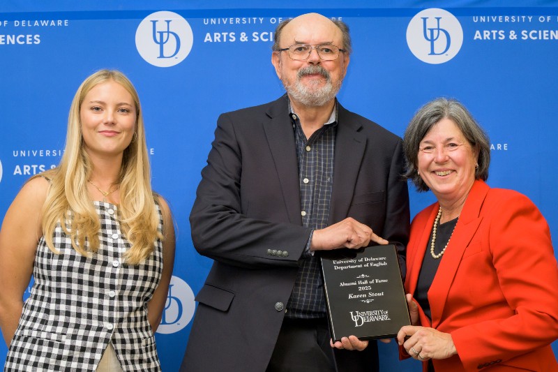 Three smiling people standing in front of a blue background with the University of Delaware logo with the man  and woman on right holding an award plaque.