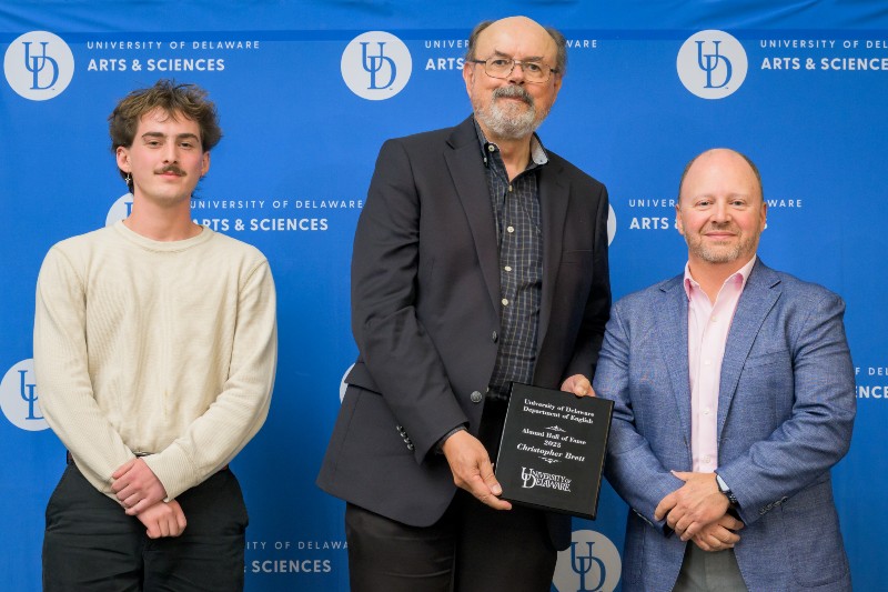 Three smiling people standing in front of a blue background with the University of Delaware logo with the two men on right holding an award plaque.