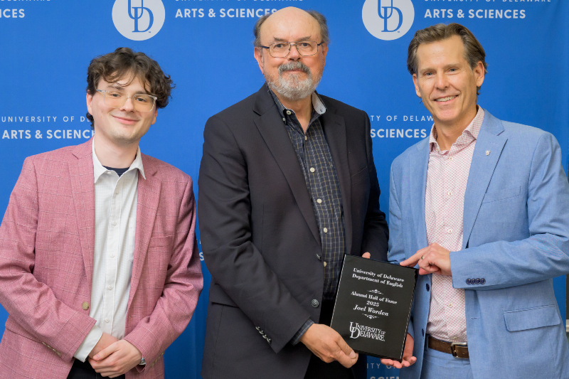 Three smiling people standing in front of a blue background with the University of Delaware logo with the two men on right holding an award plaque.