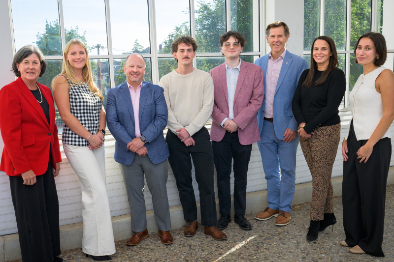 A group of eight smiling people standing in front of a sunny window.