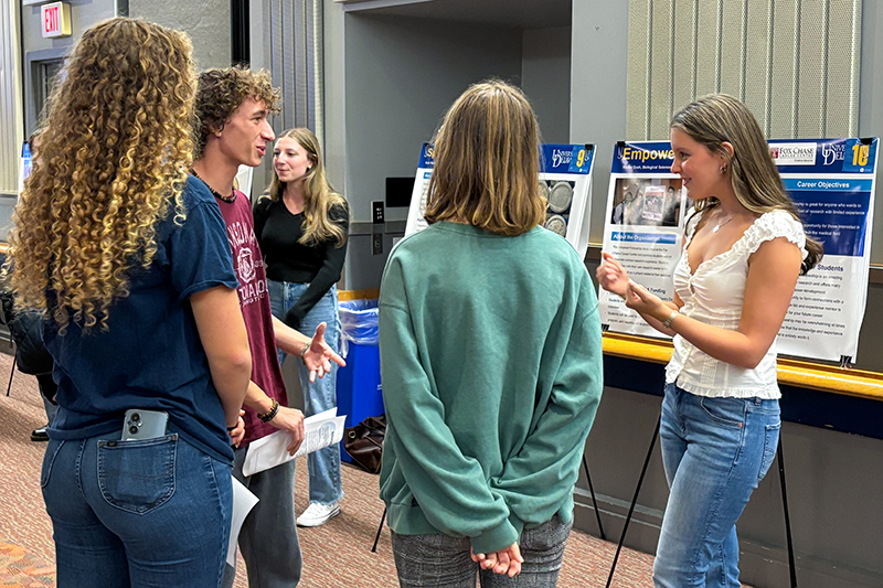 Rachel Dash Empower Fellowship Fox Chase Cancer Center A student at a student center stands next to posters on easels and talks to a group of three college students.