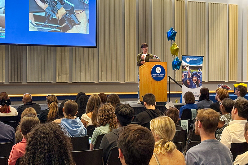 UD internship symposium lightning talk Student Success Center A college student standing behind a podium on a stage inside a student center delivers a presentation to a large audience.