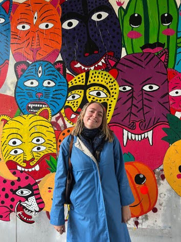 A college student in a blue raincoat in front of a colorful mural of characters.