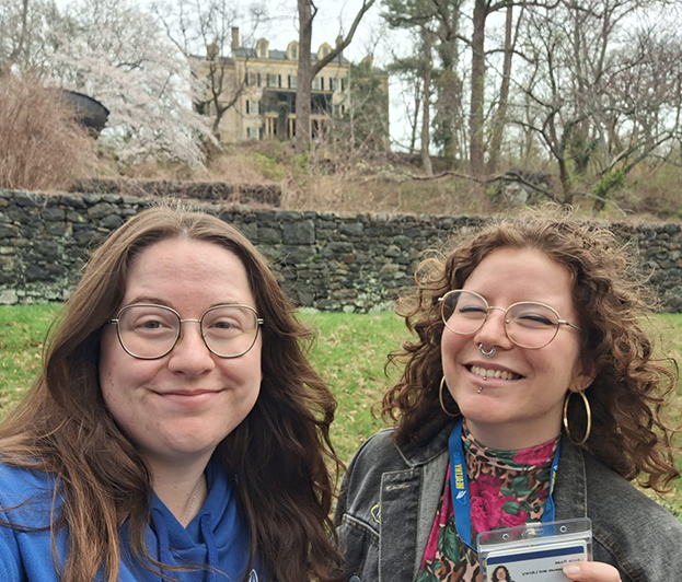 Two UD graduate students standing outside in a wooded area with a stone wall and historical building in the background.