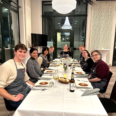 Eight college students wearing gray aprons sit down for dinner at a long table.