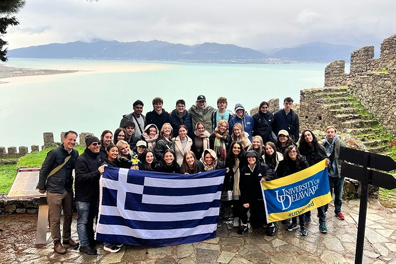 A group of smiling students and chaperones gather for a group photo in front of the blue Mediterranean Sea while holding University of Delaware and Greek flags.