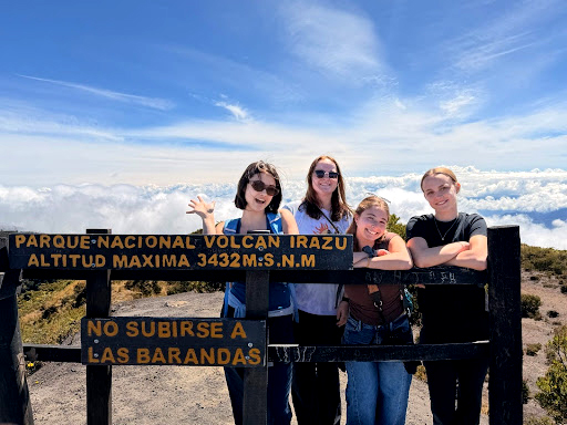 Four college students on a mountain top stand next to a sign for the Irazú Volcano National Park on a sunny day.