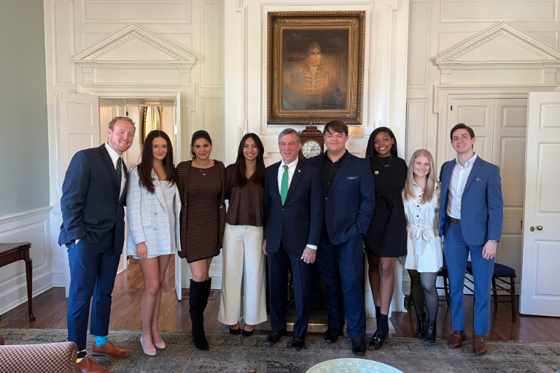 John Carney stands in the middle of a group of eight formally dressed college students gathered for a group photo inside the Delaware Governor's Mansion.