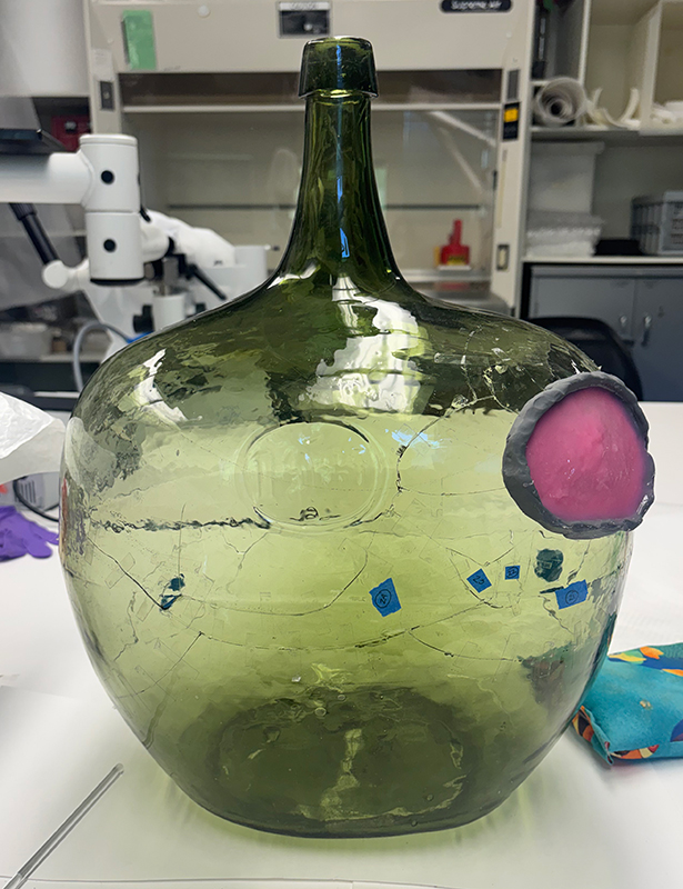 A reassembled green glass jug with blue tape and a oval portion of pink silicone on one side.